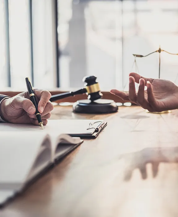 Two people at a sunny table; one writes on a document while a gavel sits between them.